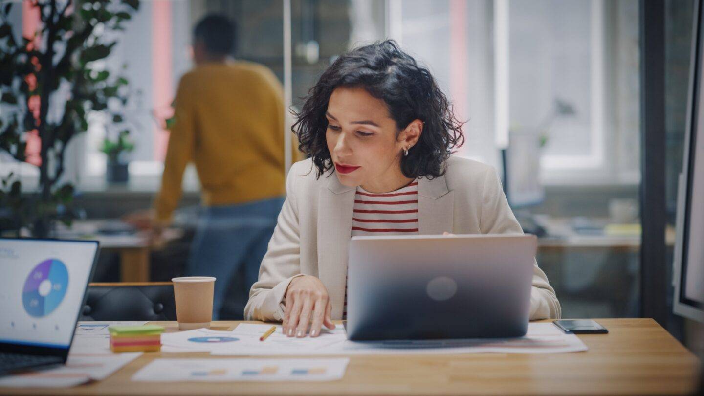 Female professional working at a laptop and looking at graphs on a document